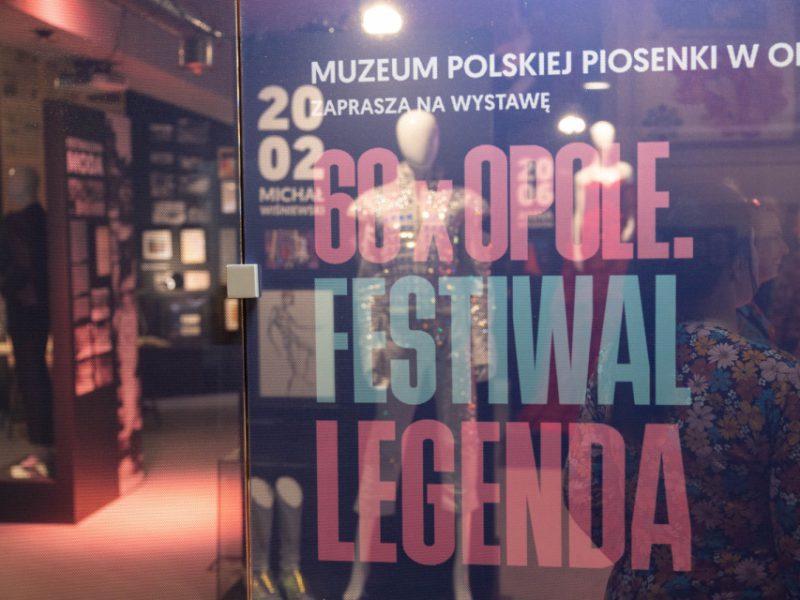 Wejście na salę wystawy, na szklanej ścianie widoczny plakat z wydarzenia, w głębi sala z wystawą- fragment wystawy pt