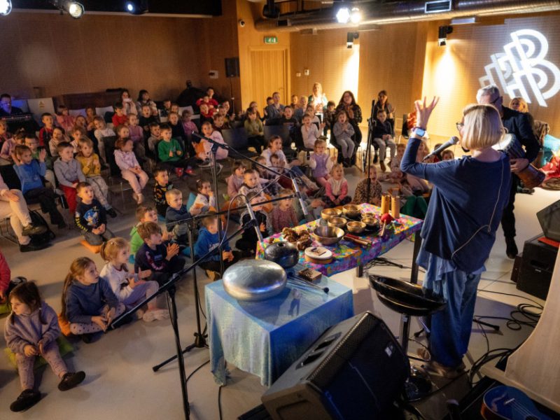 Sala muzeum, na stenie artystka z instrumentami, w tle dzieci uczestniczące w zajęciach