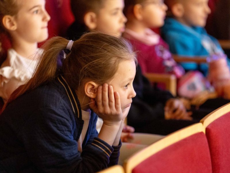 Dzieci siedzą na widowni w czerwonych fotelach, skupione i zasłuchane w to, co dzieje się na scenie. Na pierwszym planie dziewczynka w granatowej bluzie opiera głowę na dłoniach i z uwagą patrzy do przodu