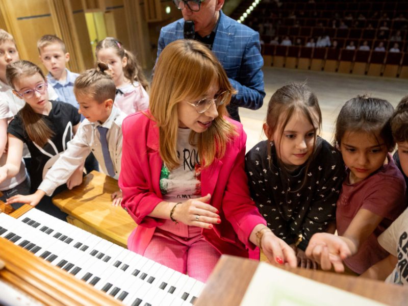 Grupa dzieci otacza kobietę ubraną w różowy garnitur, siedzącą przy dużych organach. Dzieci z zainteresowaniem przyglądają się instrumentowi i dotykają klawiszy. W tle widać pustą część widowni