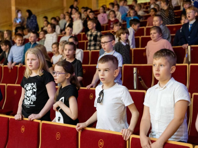 Dzieci stoją w rzędach czerwonych foteli na widowni sali koncertowej. Większość ma poważne miny, niektóre śpiewają lub mówią. W tle widoczni są inni uczniowie i opiekunowie