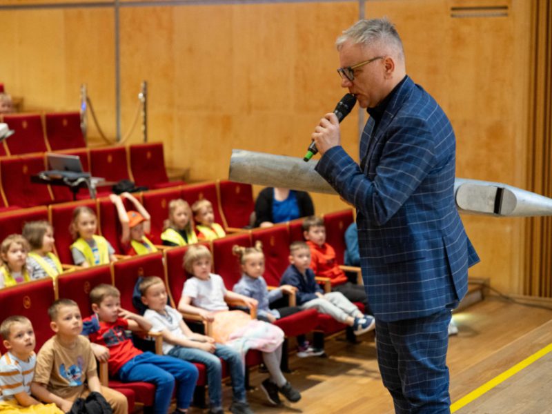 Mężczyzna w niebieskiej marynarce w kratę prowadzi spotkanie w sali koncertowej, trzymając w ręku mikrofon oraz duży element piszczałki organowej. Przed nim, w czerwonych fotelach, siedzi grupa dzieci w wieku szkolnym, uważnie słuchających