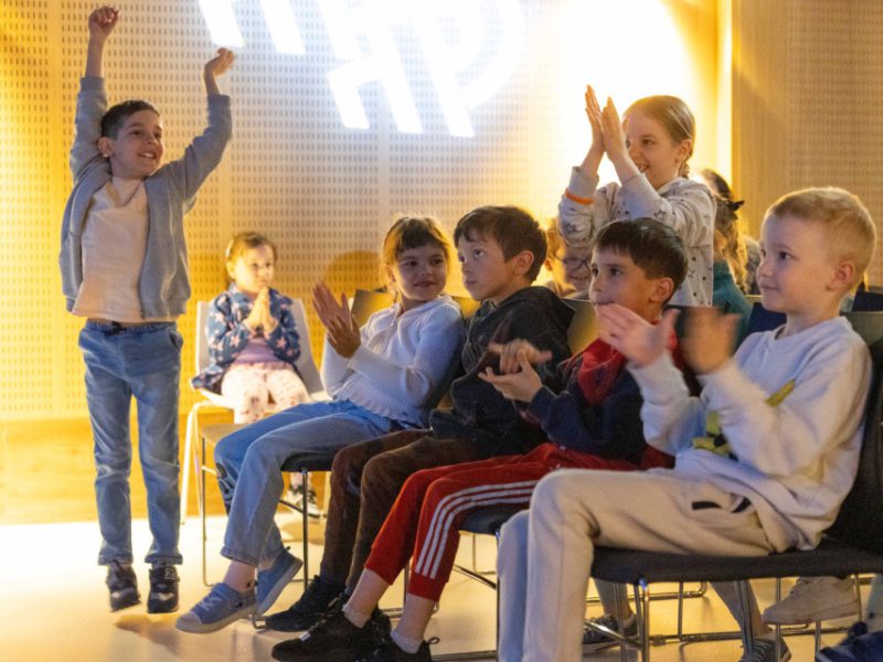 Grupa dzieci siedzi na krzesłach w sali, oglądając występ. Niektóre dzieci klaszczą, inne uśmiechają się i okazują entuzjazm. Dziewczynka stojąca po lewej stronie podnosi ręce w górę, wyrażając radość i zaangażowanie