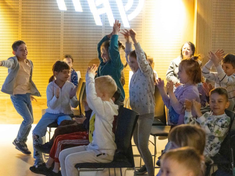 Grupa dzieci w sali, entuzjastycznie reagujących na występ – część z nich stoi, podnosi ręce w górę i bije brawo, inne siedzą i klaszczą. W tle widoczna dorosła osoba uśmiechająca się do dzieci