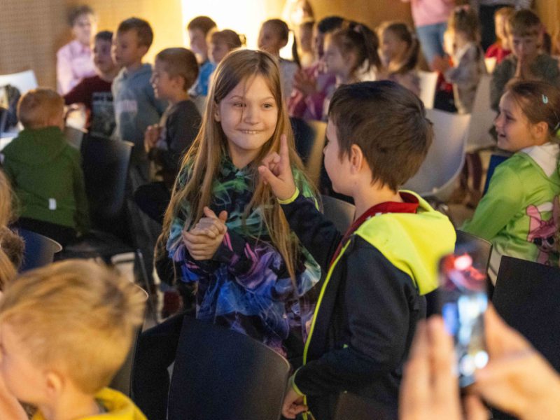 Dziewczynka i chłopiec w kolorowych bluzach uśmiechają się i rozmawiają ze sobą wśród grupy dzieci siedzących na krzesłach. W tle widać inne dzieci, które także uczestniczą w wydarzeniu