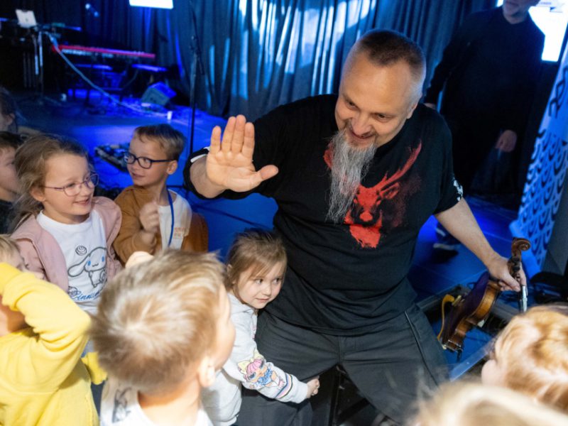Mężczyzna z długą, siwą brodą uśmiecha się i przybija piątkę z grupą dzieci. W drugiej ręce trzyma skrzypce. Dzieci są radosne i otaczają go z zainteresowaniem