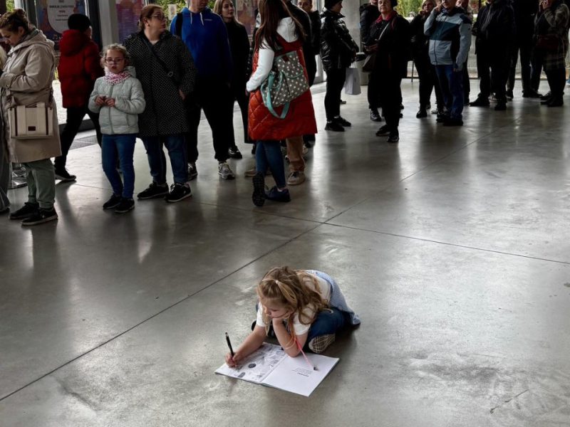 W przestronnym, przeszklonym holu budynku stoi długa kolejka ludzi ubranych w kurtki i płaszcze. Na pierwszym planie, na podłodze z jasnego betonu, mała dziewczynka w jasnej koszulce i ogrodniczkach leży na brzuchu i rysuje w dużym zeszycie lub kolorowance, ignorując oczekiwanie innych. W tle widać drzewa za szybami