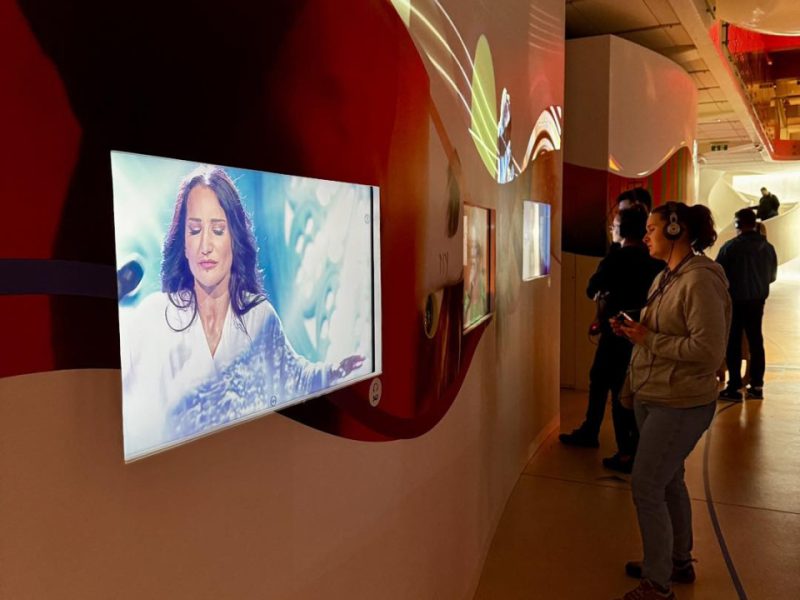 Wnętrze muzeum z interaktywną ekspozycją multimedialną. Na ścianie wyświetlany jest obraz kobiety o długich ciemnych włosach, ubranej na biało, z zamkniętymi oczami, w tle abstrakcyjne wzory. Po prawej stronie osoba w słuchawkach korzysta z urządzenia audioprzewodnika, oglądając ekspozycję