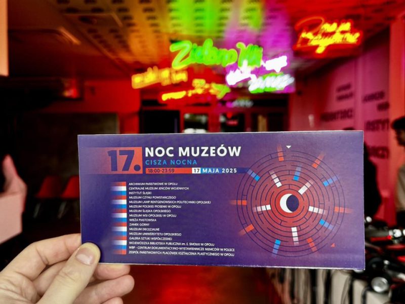 Ulotka trzymana w dłoni na tle wnętrza z kolorowymi neonami. Na ulotce granatowo-fioletowe tło z czerwonymi i niebieskimi elementami graficznymi. Duży napis: „17. Noc Muzeów – Cisza Nocna, 18:00–23:59, 17 maja 2025”. Po lewej stronie lista uczestniczących instytucji, m.in. Archiwum Państwowe w Opolu, Muzeum Polskiej Piosenki, Muzeum Śląska Opolskiego, Zamek Górny, Galeria Sztuki Współczesnej. Po prawej okrągły abstrakcyjny wzór z półksiężycem w środku