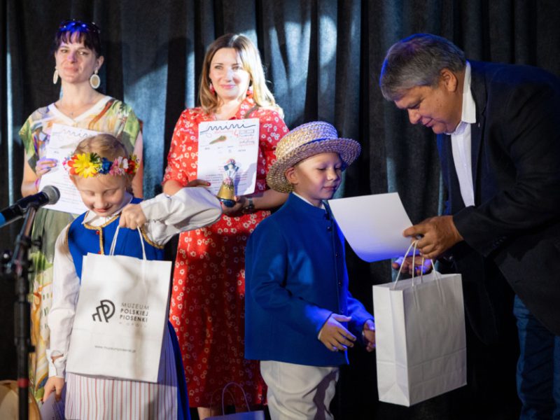Na scenie podczas uroczystości dzieci w strojach ludowych odbierają nagrody. Dziewczynka w wianku z kwiatów i chłopiec w niebieskiej marynarce oraz słomkowym kapeluszu trzymają torby z logo „Muzeum Polskiej Piosenki”. Obok nich stoją dwie kobiety z dyplomami i mężczyzna w marynarce, który wręcza dzieciom dokument i upominek