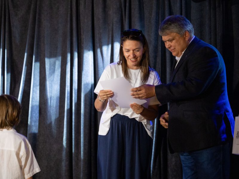 Mężczyzna w ciemnej marynarce wręcza dokument uśmiechniętej kobiecie w białej bluzce i granatowej spódnicy. Kobieta trzyma kartkę i spogląda na nią z radością. Po lewej stronie kadru stoi chłopiec w białej koszuli, patrzący w stronę dorosłych. W tle czarna zasłona
