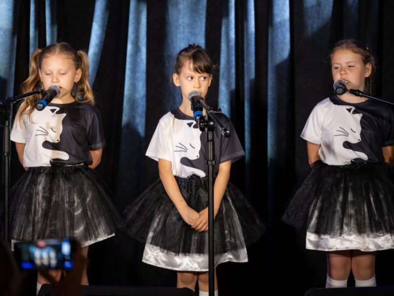 Trzy dziewczynki w identycznych strojach scenicznych – czarne tiulowe spódniczki i koszulki z motywem kota – stoją obok siebie na scenie. Każda z nich ma przed sobą mikrofon. Dziewczynka po lewej mówi do mikrofonu, środkowa stoi ze złożonymi dłońmi i patrzy na publiczność, a dziewczynka po prawej śpiewa lub recytuje