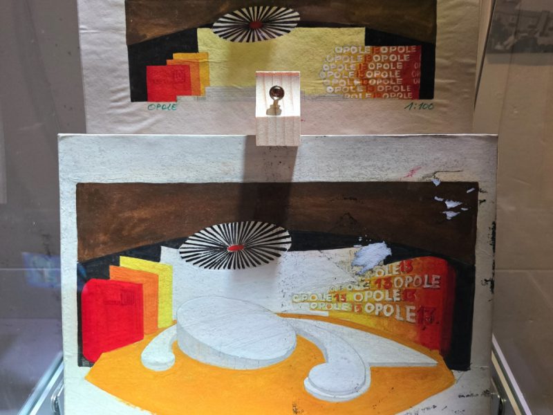 W gablocie muzealnej znajdują się dwa projekty scenografii w formie kolorowych makiet na papierze, przedstawiające scenę festiwalu w Opolu. Kompozycja zawiera geometryczne kształty w odcieniach czerwieni, żółci, brązu i bieli, z centralnie umieszczonym okrągłym podestem oraz napisem „OPOLE” powielanym na czerwonym tle. Poniżej ustawiono kilka miniaturowych plansz z plakatami i grafikami promującymi festiwal, opisanych numerami