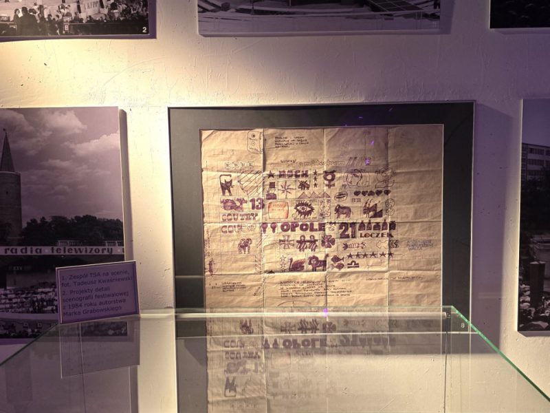 Fotografia przedstawia muzealną gablotę z dokumentami i projektami związanymi z Krajowym Festiwalem Piosenki Polskiej w Opolu. Na ścianie powyżej widoczne są czarno-białe zdjęcia sceny festiwalowej z różnych lat. W centralnym punkcie znajduje się duży arkusz papieru z odręcznymi rysunkami, napisami i symbolami – projekt scenograficzny. W gablocie pod nim ułożone są plakaty i materiały graficzne, m.in. z napisami „OPOLE ’84”, „COUNTRY”, „ROCK”