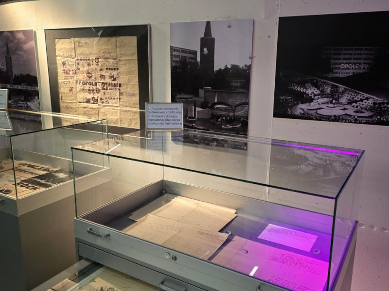 Ekspozycja muzealna poświęcona historii festiwalu w Opolu. Na ścianie wiszą archiwalne czarno-białe fotografie sceny i występów artystów. Pod nimi stoją szklane gabloty z projektami scenografii, dokumentami i rysunkami technicznymi. W jednej z szuflad widoczny jest duży projekt dekoracji koncertowej, a na kartkach w gablocie znajdują się szczegółowe szkice i opisy techniczne
