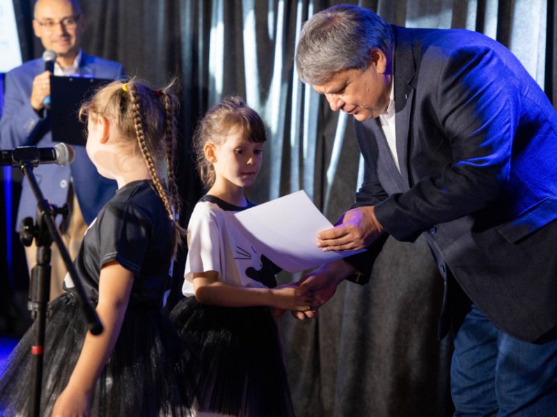 Mężczyzna w garniturze wręcza dyplom dwóm małym dziewczynkom stojącym na scenie. Dziewczynki ubrane są w białe koszulki i czarne spódniczki. W tle widać drugiego mężczyznę z mikrofonem i teczką, zapowiadającego wydarzenie