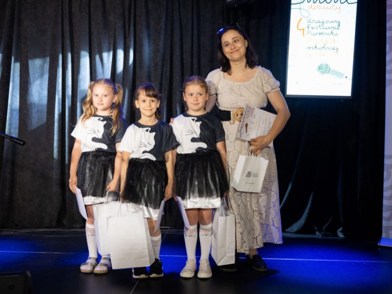 Trzy dziewczynki w białych koszulkach z czarnym nadrukiem kota i czarnych tiulowych spódniczkach stoją na scenie obok kobiety w jasnej koronkowej sukience. Wszystkie trzymają w rękach białe torby z upominkami i dyplomy. W tle widoczny ekran z napisem „Krajowy Festiwal Piosenki Dziecięcej i Młodzieżowej” oraz grafiką mikrofonu