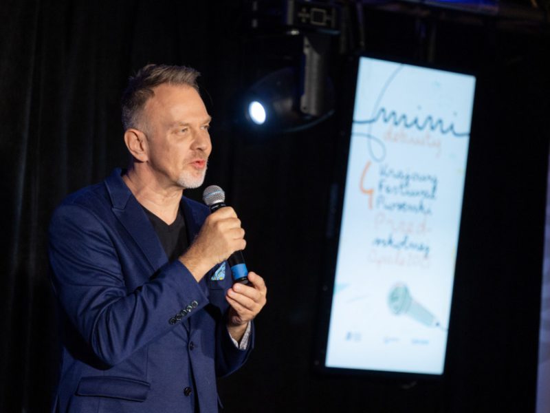 Mężczyzna w granatowej marynarce przemawia do mikrofonu na scenie. W tle widoczny jest ekran z napisem „Mini Debiuty 4. Krajowy Festiwal Piosenki Przedszkolnej” oraz rysunkiem mikrofonu