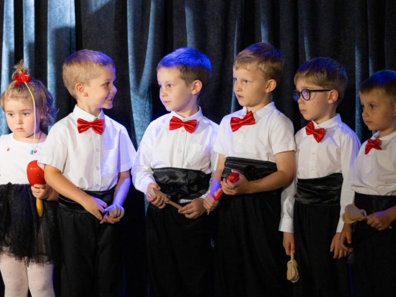 Grupa dzieci stoi na scenie na tle ciemnej kotary. Dziewczynka z lewej strony trzyma czerwoną grzechotkę, a obok niej kilku chłopców ubranych w białe koszule z czerwonymi muszkami i czarnymi spodniami trzyma instrumenty perkusyjne, m.in. kastaniety i pałeczki. Dzieci wyglądają na skupione i przygotowane do występu