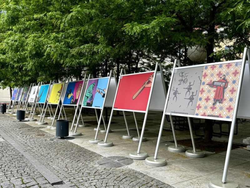 Plenerowa wystawa plakatów ustawionych w szeregu na metalowych stojakach, na tle zielonych drzew. Każdy plakat ma intensywne, jednolite tło w różnych kolorach i przedstawia minimalistyczne ilustracje związane z muzyką i kulturą