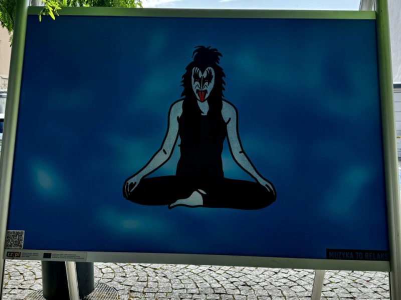 Plansza wystawowa z ilustracją postaci w makijażu scenicznym w stylu zespołu KISS, siedzącej po turecku w pozycji medytacyjnej, na niebieskim tle. Podpis: „MUZYKA TO RELAKS”