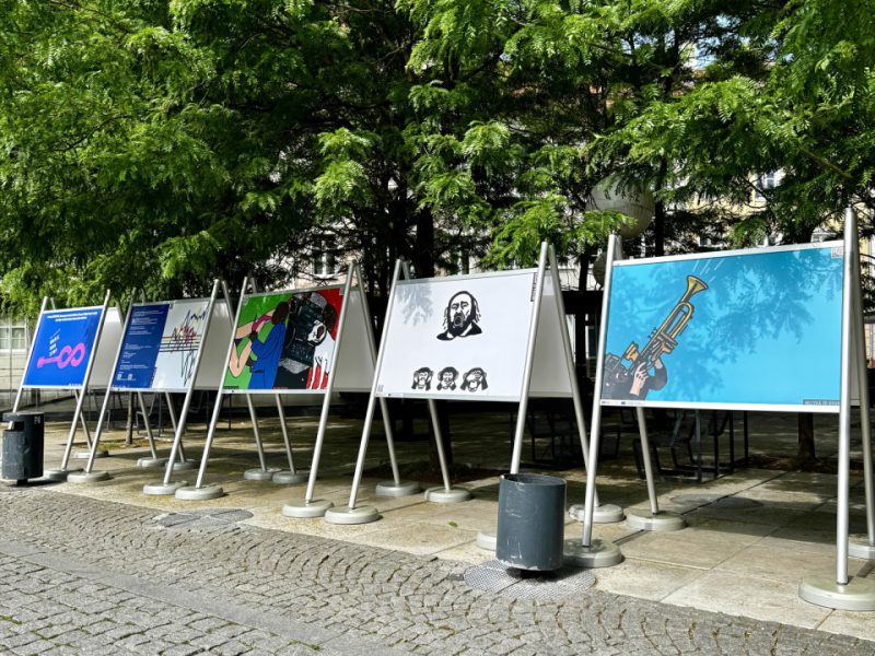 Szereg kolorowych plansz ustawionych w metalowych ramach na chodniku wzdłuż rzędu drzew. Każda plansza przedstawia inną ilustrację na tle jednolitego koloru, m.in. postacie, symbole i instrumenty muzyczne