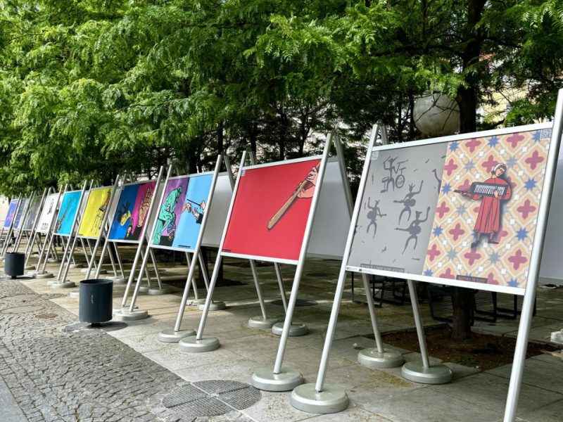 Ulica wyłożona kostką brukową z rzędem metalowych stojaków prezentujących plakaty z grafikami artystycznymi. W tle drzewa o gęstych, zielonych liściach oraz fragmenty miejskiej zabudowy