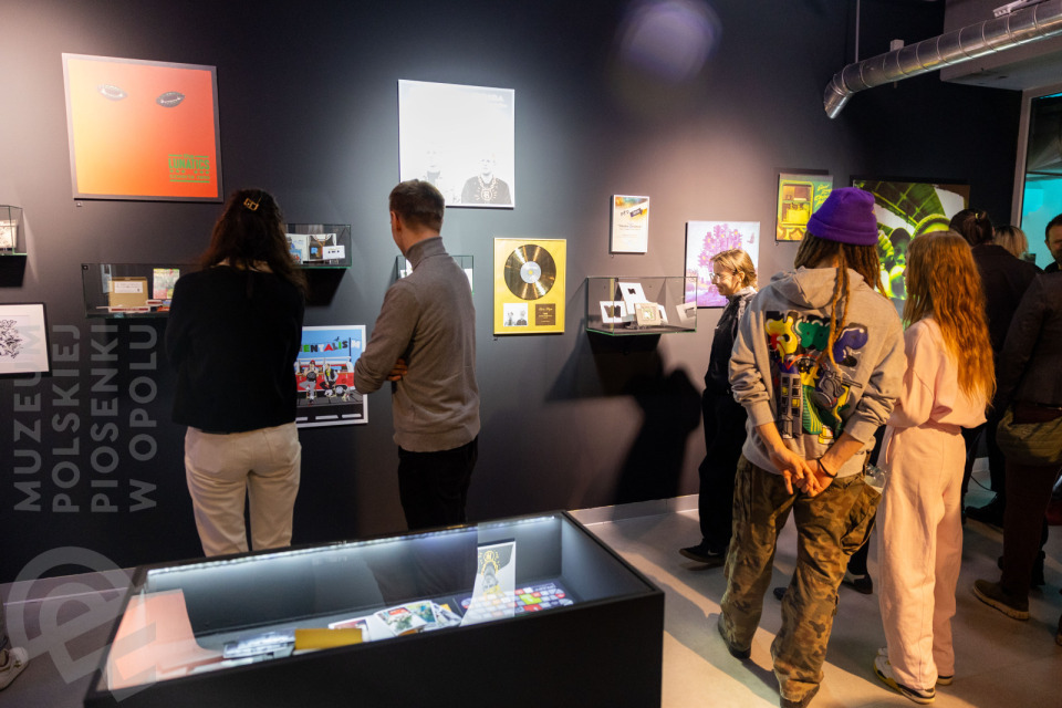 Wnętrze Muzeum Polskiej Piosenki w Opolu. Kilka osób ogląda eksponaty zawieszone na ciemnej ścianie — wśród nich okładki płyt, złota płyta, fotografie i grafiki. Na pierwszym planie, po lewej stronie, przeszklona gablota z płytami CD i innymi przedmiotami kolekcjonerskimi