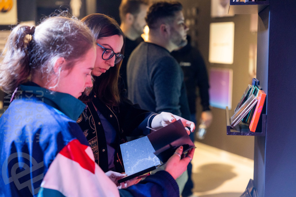Dwie młode kobiety stoją przy półce w muzeum i oglądają opakowanie płyty CD. Na półce obok widoczny rząd kolorowych pudełek z płytami. W tle inni zwiedzający obserwują eksponaty na ścianach