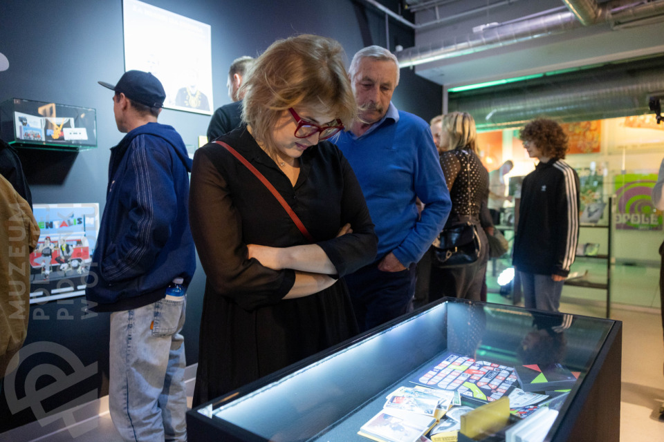 Kobieta w czarnej sukience i okularach o czerwonych oprawkach pochyla się nad podświetloną gablotą z eksponatami w Muzeum Polskiej Piosenki w Opolu. Obok niej starszy mężczyzna w niebieskim swetrze, w tle inni zwiedzający oglądają wystawę