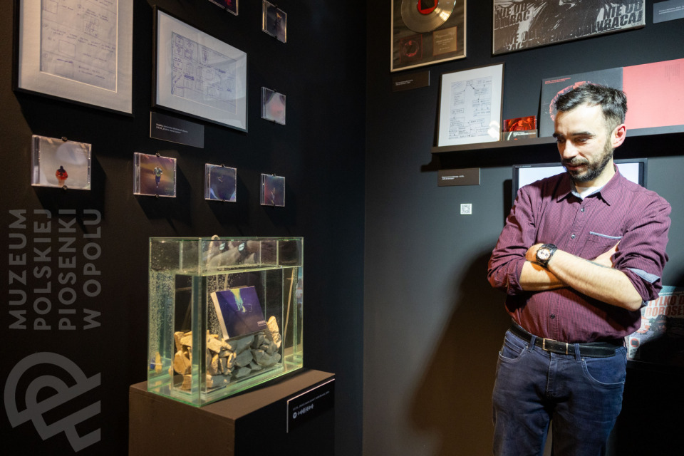 Mężczyzna w bordowej koszuli stoi z założonymi rękami przed ekspozycją w muzeum, obejmującą szkice, fotografie w małych ramkach oraz akwarium wypełnione kamieniami i płytą CD, umieszczone na postumencie