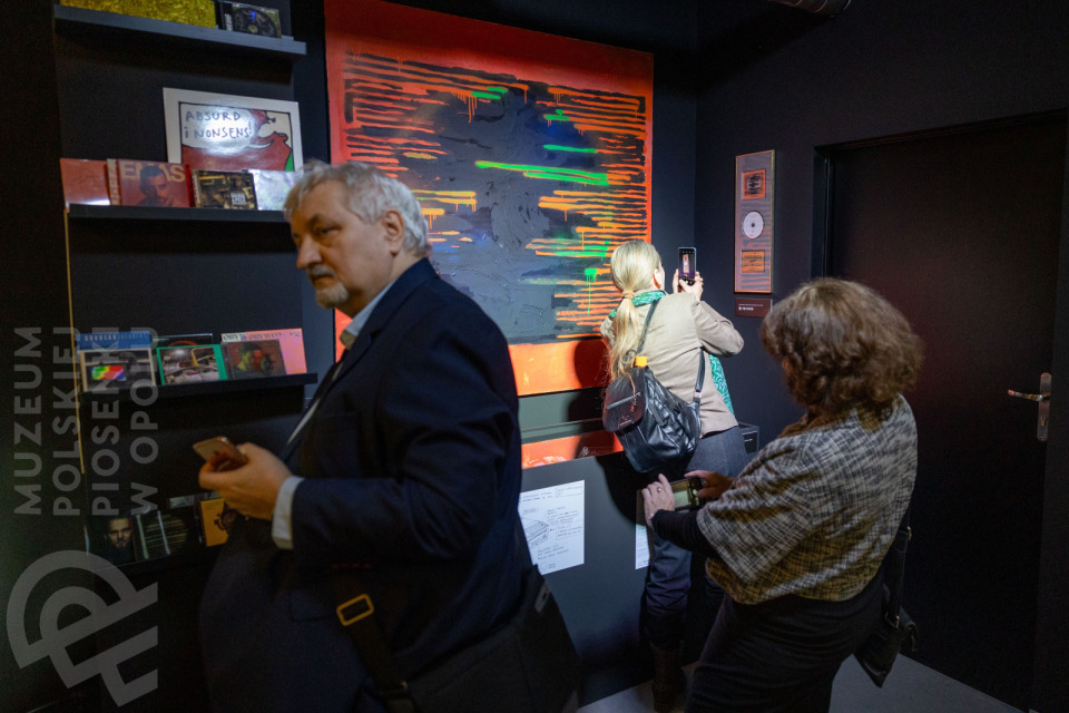 Trzy osoby w sali wystawowej Muzeum Polskiej Piosenki w Opolu. Na ścianie widoczny duży, abstrakcyjny obraz w odcieniach szarości, pomarańczu i zieleni. Kobieta w jasnej kurtce fotografuje dzieło telefonem, obok niej stoi mężczyzna w garniturze oraz kobieta w kraciastej narzutce oglądająca ekspozycję. Na półkach po lewej znajdują się płyty CD