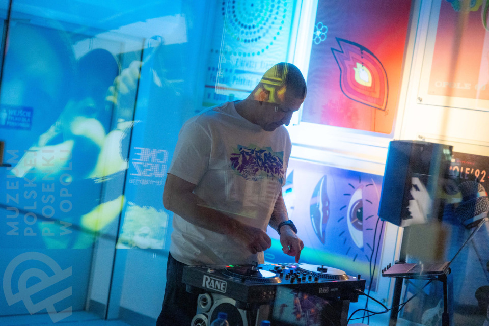 DJ w białej koszulce z fioletowym nadrukiem miksuje muzykę na konsoli w przestrzeni wystawowej Muzeum Polskiej Piosenki w Opolu. W tle barwnie oświetlone plakaty muzyczne w odcieniach niebieskiego, czerwonego i pomarańczowego