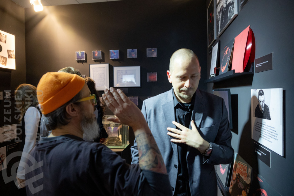 Rozmowa dwóch mężczyzn w sali wystawowej muzeum. Mężczyzna po lewej, w pomarańczowej czapce beanie i okularach, gestykuluje, zwracając się do mężczyzny po prawej, ubranego w elegancki, ciemnoszary garnitur. W tle na czarnej ścianie wiszą winyle, zdjęcia i gabloty z eksponatami