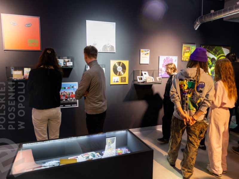 Wnętrze Muzeum Polskiej Piosenki w Opolu. Kilka osób ogląda eksponaty zawieszone na ciemnej ścianie — wśród nich okładki płyt, złota płyta, fotografie i grafiki. Na pierwszym planie, po lewej stronie, przeszklona gablota z płytami CD i innymi przedmiotami kolekcjonerskimi