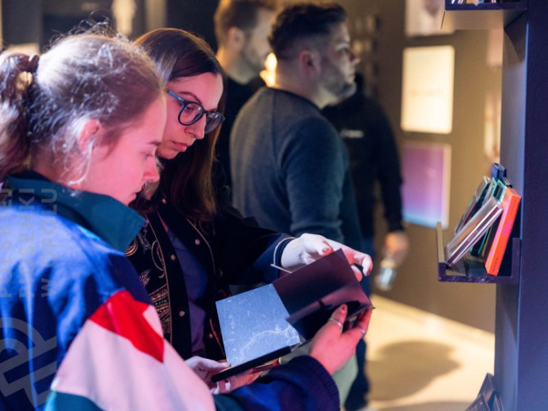 Dwie młode kobiety stoją przy półce w muzeum i oglądają opakowanie płyty CD. Na półce obok widoczny rząd kolorowych pudełek z płytami. W tle inni zwiedzający obserwują eksponaty na ścianach