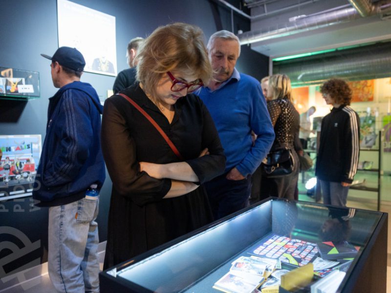 Kobieta w czarnej sukience i okularach o czerwonych oprawkach pochyla się nad podświetloną gablotą z eksponatami w Muzeum Polskiej Piosenki w Opolu. Obok niej starszy mężczyzna w niebieskim swetrze, w tle inni zwiedzający oglądają wystawę