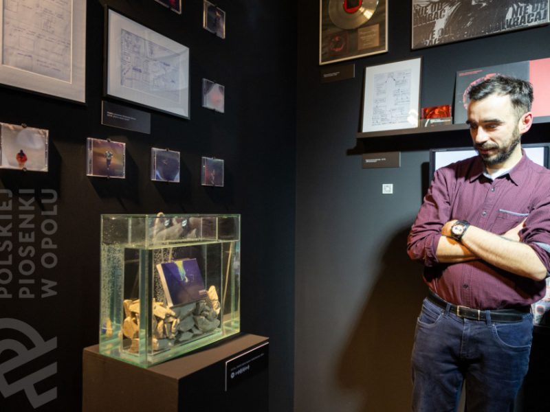 Mężczyzna w bordowej koszuli stoi z założonymi rękami przed ekspozycją w muzeum, obejmującą szkice, fotografie w małych ramkach oraz akwarium wypełnione kamieniami i płytą CD, umieszczone na postumencie