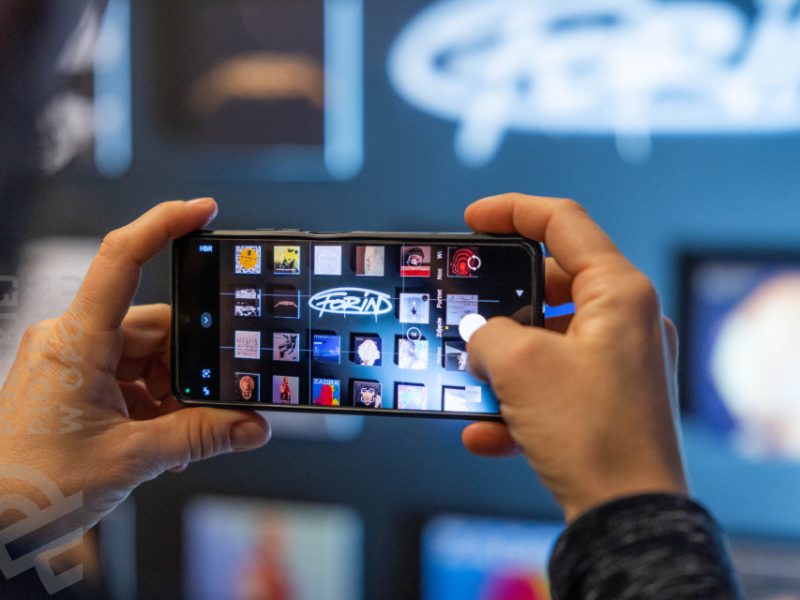 Zbliżenie na dłonie osoby fotografującej smartfonem ścianę z okładkami albumów muzycznych, widocznych na ekranie telefonu oraz w tle