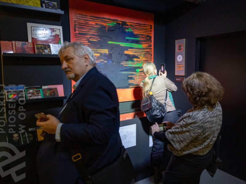 Trzy osoby w sali wystawowej Muzeum Polskiej Piosenki w Opolu. Na ścianie widoczny duży, abstrakcyjny obraz w odcieniach szarości, pomarańczu i zieleni. Kobieta w jasnej kurtce fotografuje dzieło telefonem, obok niej stoi mężczyzna w garniturze oraz kobieta w kraciastej narzutce oglądająca ekspozycję. Na półkach po lewej znajdują się płyty CD
