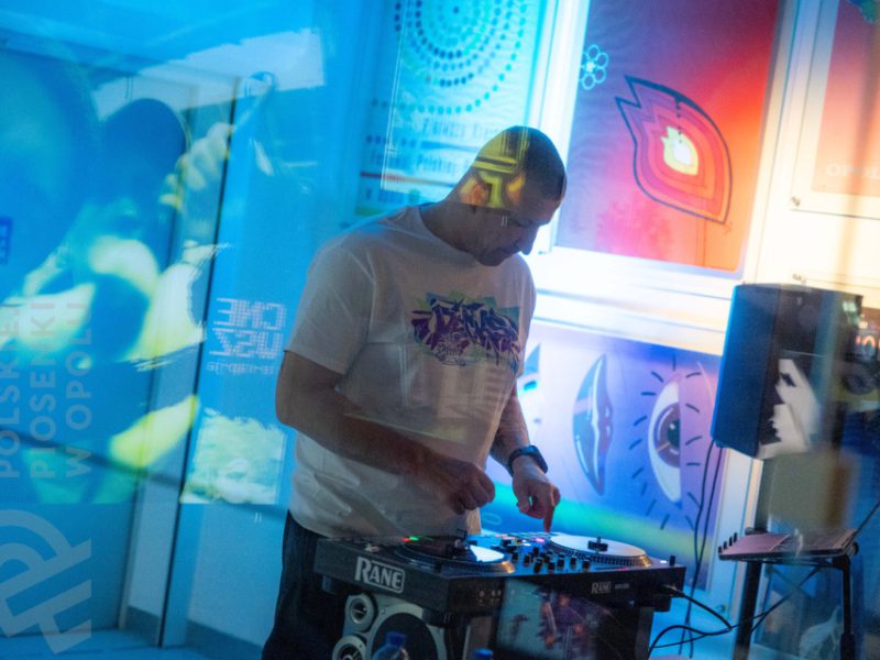 DJ w białej koszulce z fioletowym nadrukiem miksuje muzykę na konsoli w przestrzeni wystawowej Muzeum Polskiej Piosenki w Opolu. W tle barwnie oświetlone plakaty muzyczne w odcieniach niebieskiego, czerwonego i pomarańczowego