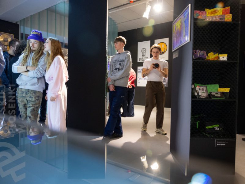 Wnętrze muzeum z grupą młodych ludzi oglądających wystawę. Po lewej para – mężczyzna w czapce z daszkiem i długich dredach oraz kobieta w jasnoróżowym stroju – patrzy na eksponaty. W tle inni zwiedzający, a po prawej widać półki z kolorowymi przedmiotami i grafikę na ścianie