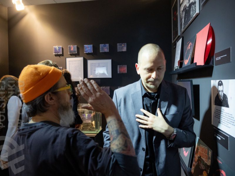 Rozmowa dwóch mężczyzn w sali wystawowej muzeum. Mężczyzna po lewej, w pomarańczowej czapce beanie i okularach, gestykuluje, zwracając się do mężczyzny po prawej, ubranego w elegancki, ciemnoszary garnitur. W tle na czarnej ścianie wiszą winyle, zdjęcia i gabloty z eksponatami