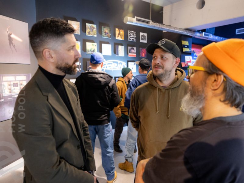 Trzech mężczyzn w rozmowie w sali wystawowej. Po lewej mężczyzna w ciemnej marynarce i czarnym golfie, pośrodku mężczyzna w brązowej bluzie z kapturem i czapce z daszkiem, po prawej mężczyzna w pomarańczowej czapce i żółtych okularach. W tle na ścianie kolorowe okładki płyt i grafiki