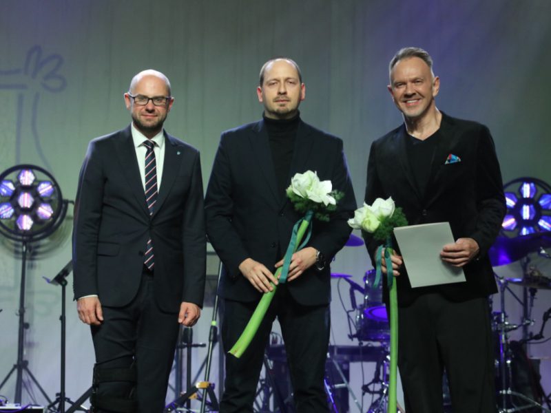 Trzej mężczyźni w eleganckich garniturach stoją na scenie, dwaj z nich trzymają długie łodygi białych kwiatów, a jeden dodatkowo kartkę; za nimi widoczne są reflektory sceniczne i zestaw perkusyjny