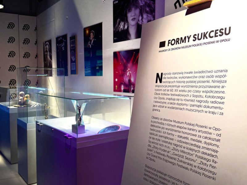 Fragment wystawy „Formy sukcesu” w Muzeum Polskiej Piosenki w Opolu – po prawej duża plansza z tekstem wprowadzającym, po lewej rząd szklanych gablot z nagrodami, a na ścianie powyżej kolorowe portrety polskich wykonawców w fioletowo-niebieskim świetle