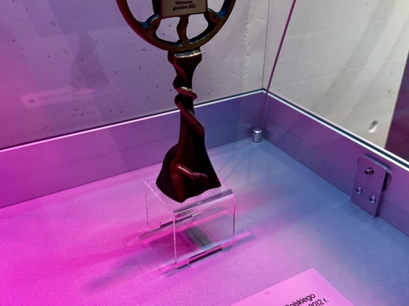 Pojedyncza statuetka „Honorowy Złoty Mikrofon” w gablocie – smukła, metalowa forma przypominająca stary mikrofon na podstawce, przed nią biała tabliczka z opisem nagrody dla Grażyny Łobaszewskiej