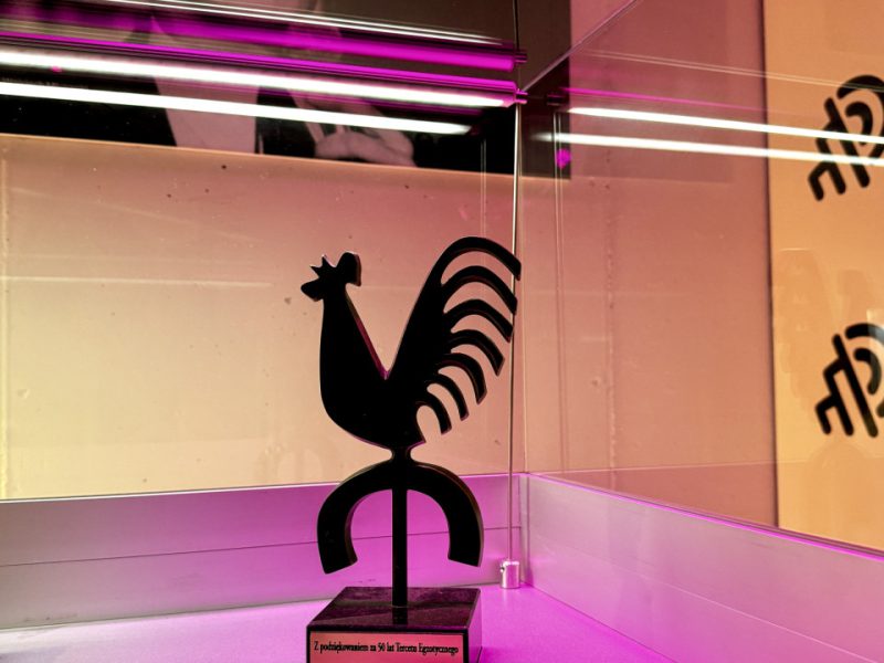 Czarna, graficzna statuetka w kształcie koguta na podstawce w szklanej gablocie; poniżej opis nagrody dla Tercetu Egzotycznego, a w tle na ścianie duży czarno-biały portret śpiewającego artysty odbijający się w szybach