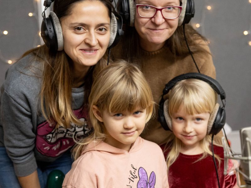 Dwie kobiety i dwie dziewczynki stoją razem w studiu nagraniowym. Dzieci trzymają instrumenty perkusyjne, a wszyscy mają na głowie duże słuchawki