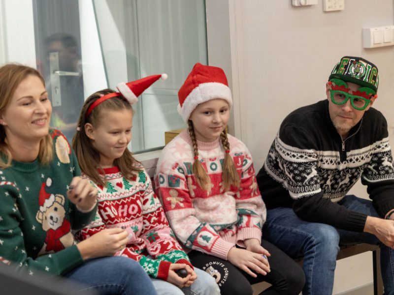 Dwie dziewczynki i dwoje dorosłych siedzą razem w studiu. Wszyscy mają na sobie świąteczne swetry i akcesoria, a jedna osoba nosi duże zielone okulary z motywem choinki